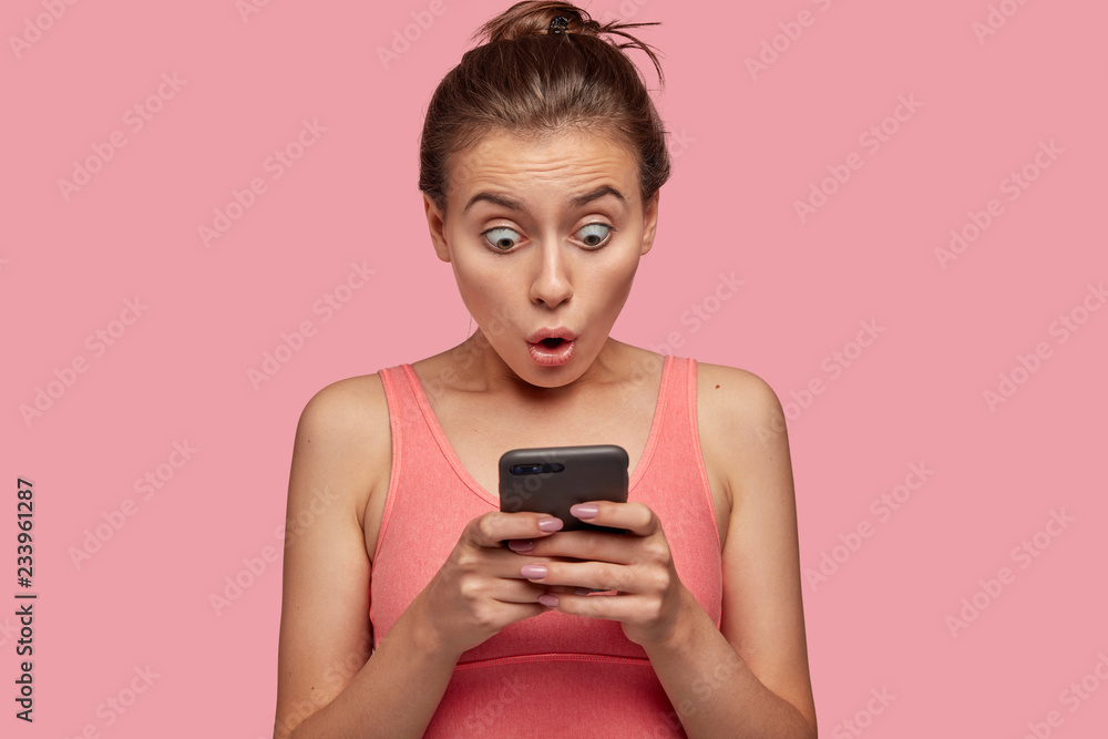 Stupefied emotional woman stares with bugged eyes at smart phone, feels shocked as reads news at web page, connected to wireless internet, has break after training with coach in gym, wears pink top Stupefied emotional woman stares with bugged eyes at smart phone, feels shocked as reads news at web page, connected to wireless internet, has break after training with coach in gym, wears pink top