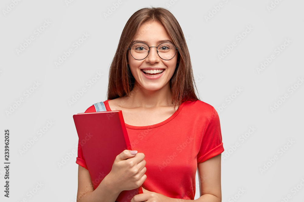 Positive Caucasian girl with charming smile, wears red t shirt, holds textbook, models against white background, has mood for studying, wears optical glasses for good vision. Youth, learning concept Positive Caucasian girl with charming smile, wears red t shirt, holds textbook, models against white background, has mood for studying, wears optical glasses for good vision. Youth, learning concept