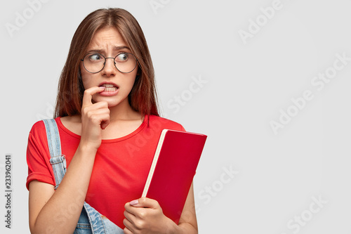 Worried anxious young woman keeps index finger near mouth, looks desperately aside wears round spectacles, carries red textbook, models against white background, free space for your promotion