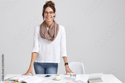 Satisfied intelligent female tutor wears white jumper and jeans, keeps hands on table, ready to give private lessons, drinks coffee, stands against white background. Woman works freelance at project photo