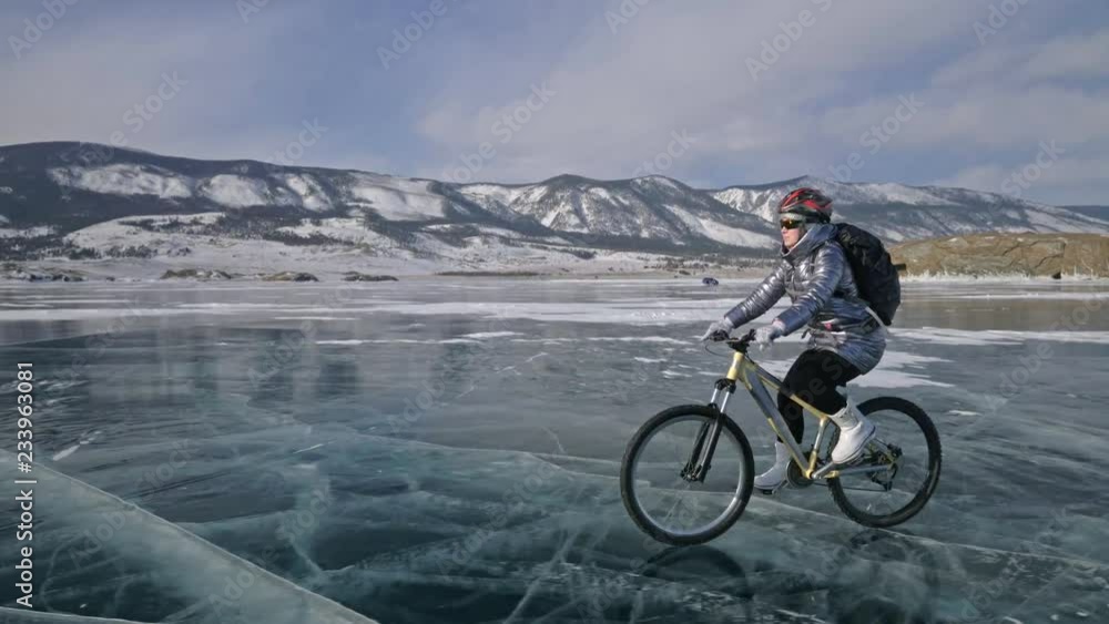 Woman is riding bicycle on the ice. The girl is dressed in a silvery down jacket, cycling backpack and helmet. Ice of the frozen Lake Baikal. The tires on the bicycle are covered with special spikes