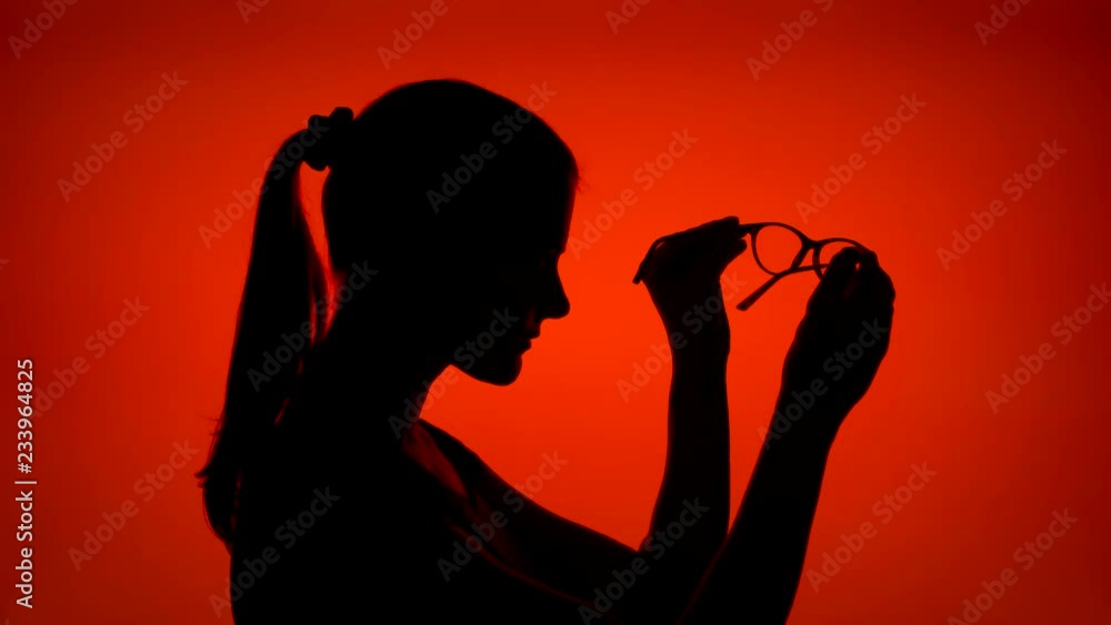 Silhouette of young woman with eyeglasses. Female's face in profile putting on glasses on red background. Black contour shadow of teenager's half-face holding spectacles Silhouette of young woman with eyeglasses. Female's face in profile putting on glasses on red background. Black contour shadow of teenager's half-face holding spectacles