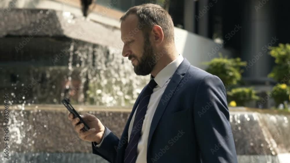 Young businessman texting on smartphone standing near fountain in city Young businessman texting on smartphone standing near fountain in city