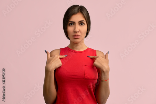 A pretty woman has a puzzled expression, raises her eyebrow, while pointing to herself, shows an empty place of clothing, posing at the pink wall. Brunette shows a place for your content
