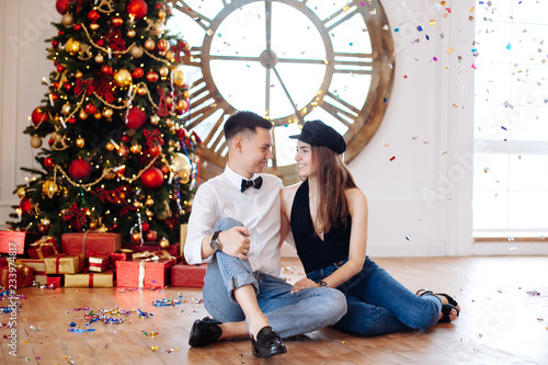 Cheerful stylish couple sitting near red Christmas tree under confetti