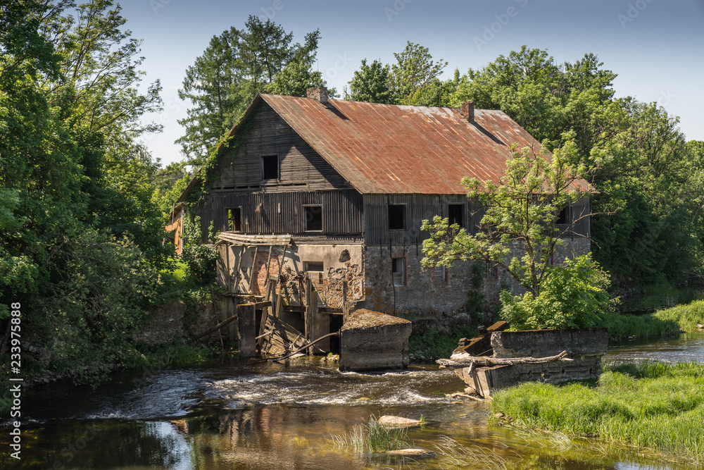 Ancient abandoned water mill surrounded by beautiful nature. House built of stone and wood, exterior walls and dilapidated bridge on river is reflection of trees and hous ruins
