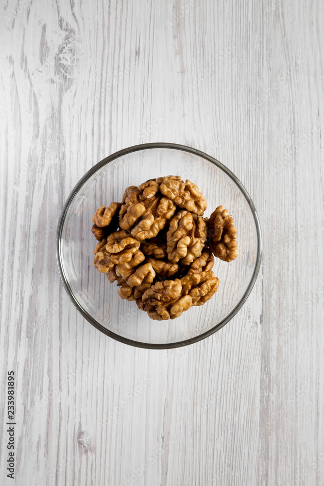 Fototapeta premium Walnuts in glass bowl over white wooden background, overhead view. Close-up. Flat lay, from above, top view.