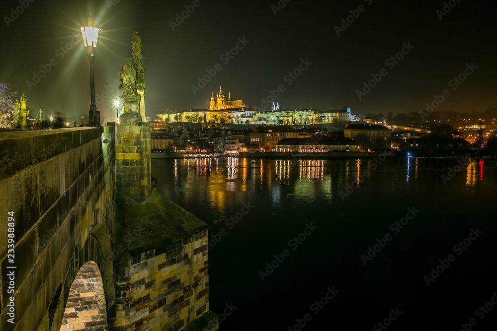 Night foggy view of famous medieval stone bridge with statues and street lights, Charles bridge, Prague castle in distance, capital of Czech Republic, colorful reflection in water of river Moldau