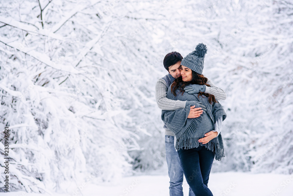 Husband in a grey sweater and jeans tenderly hugging his beautiful pregnant wife in grey knitted sweaters, hat, scarf and jeans in a snowy winter park. Pregnancy concept.