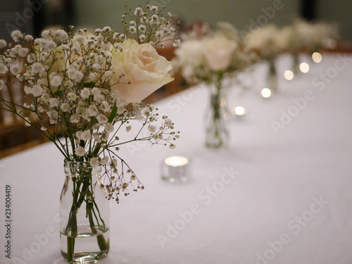 White rose and white flower with green stem in glass bottle jar vase on white table cloth with tea light candles