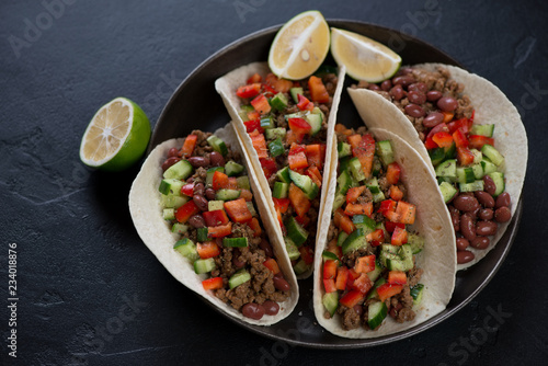 Mexican tacos with minced beef meat, red beans and fresh vegetables filling, horizontal shot over black stone background