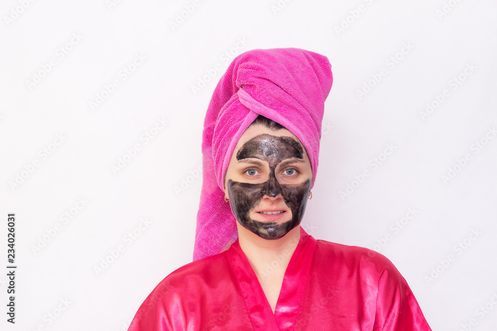 A girl on a white background with a black mask on her face against blackheads and black dots in a pink robe and a towel on her head, copy space A girl on a white background with a black mask on her face against blackheads and black dots in a pink robe and a towel on her head, copy space