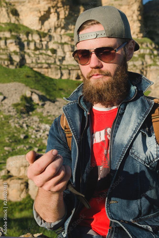 Close-up of a bearded man in jeans clothes in sunglasses and a cap with a backpack sitting at the foot of the epic rocks with a compass in his hands. Navigation travel tourism and orientation