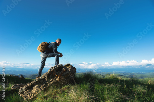Stylish bearded male traveler in sunglasses and a cap with a backpack in a denim suit and yellow shoes stands on a large stone against the background of an evening horizon. Travel and look to the