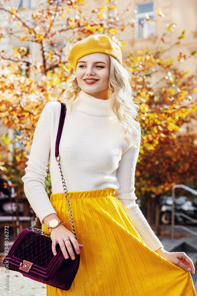 Happy smiling fashionable woman posing in autumn street, modle wearing yellow beret, white turtleneck, analog wrist watch, pleated skirt, holding purple quilted bag