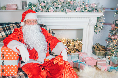Picture of man in santa claus suit sits and poses on camera. He holds one hand on present box. There are fireplace with socks and gifts behind him.