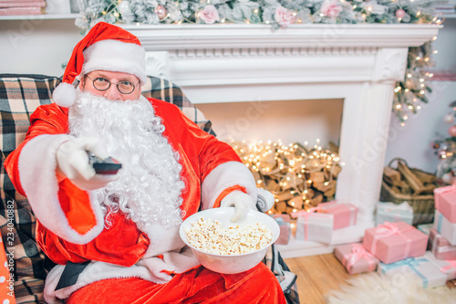 Excited Santa Claus sits at fireplace and holds remote control. Man holds bowl of popcorn. He is happy.