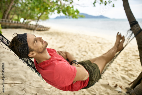 Man with stubble is lying on black hammock on sand beach