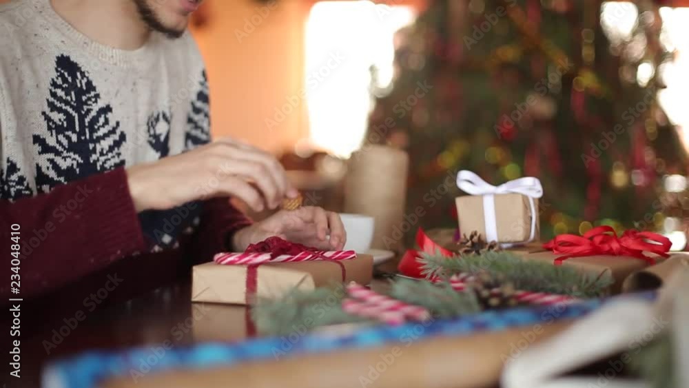 Close view of man's hands tying a bow on a gifts for Christmas. Male wrapping present boxes in paper adding fir branches, cones, cane candies on wooden table at home near New Year tree. Close view of man's hands tying a bow on a gifts for Christmas. Male wrapping present boxes in paper adding fir branches, cones, cane candies on wooden table at home near New Year tree.