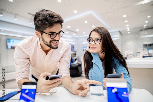 Man holding smart phone and pointing on other phone which is in woman`s hands. Tech store interior.