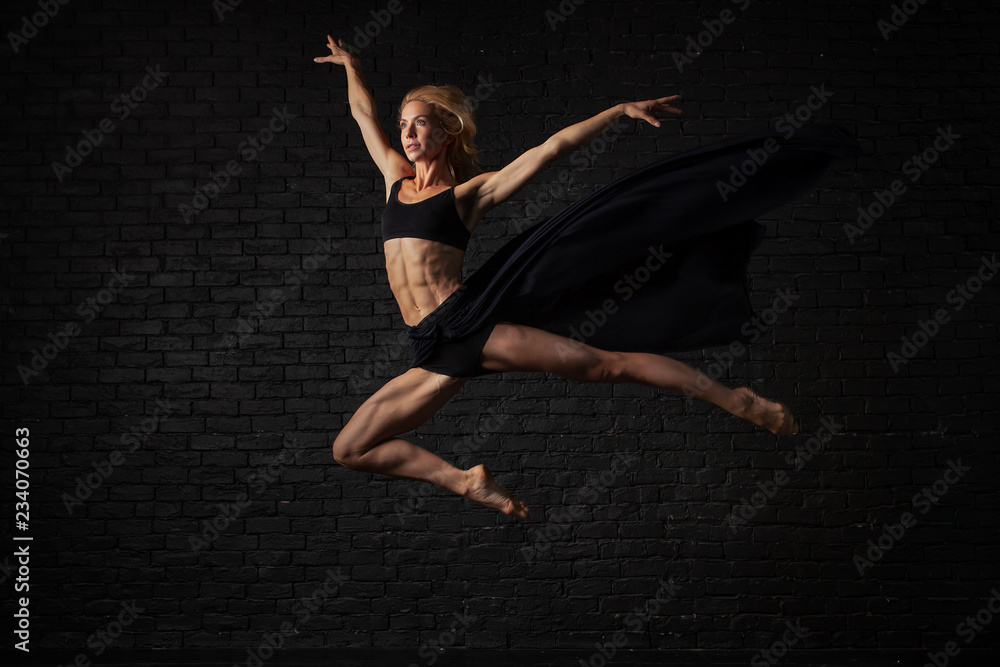 Young blonde ballerina in sportswear underwear dances and jumps in a studio with black brick on the background. modern ballet performance. Perfect fitness woman body