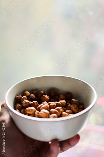 A male hand holds organic hazelnuts in a white bowl without a shell. Hazelnuts in white bowl with light blurred background. The concept of organic healthy food. Vertical, selective focus.