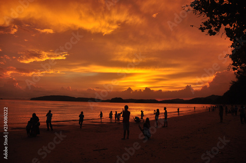 People on sandy shore in sunset, Silhouettes of people spending time on tropical coastline with golden bright sky in sunset light, Thailand.