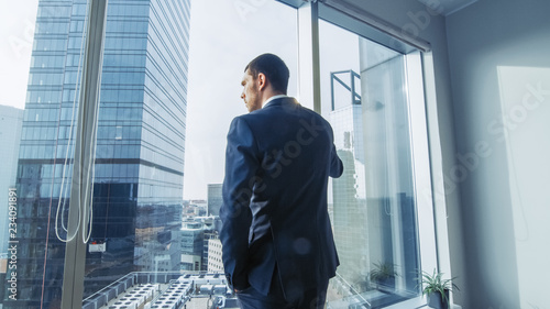 Thoughtful Businessman Wearing Suit Standing in His Office, Looking out of the Window and Contemplating Next Big Business Contract. Major City Business District with Panoramic Window View. Blue Colors photo