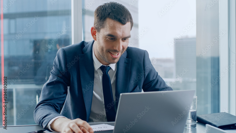 Successful Businessman Sitting at His Desk, Using His Laptop. In the Window Big City Business District View. Successful Businessman Sitting at His Desk, Using His Laptop. In the Window Big City Business District View.