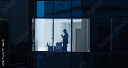 Aerial Shot: From Outside into Office Building with Businessman Using Mobile Phone and Standing by the Office Window. Beautiful Shot of The Financial Business District Skyscrapers. 