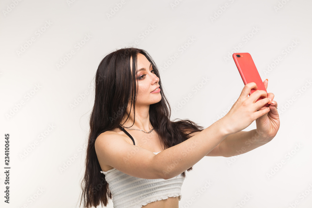 Happy european female model with dark hair enjoying indoor photoshoot. Young woman is taking a selfie on light background Happy european female model with dark hair enjoying indoor photoshoot. Young woman is taking a selfie on light background