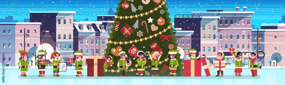 Fototapeta premium mix race elves group near decorated fir tree city building houses night winter street cityscape merry christmas happy new year concept flat horizontal banner vector illustration