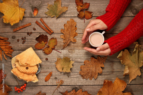 Cup of coffee with cream in hands, cinnamon sticks, waffles and autumn leaves on wooden background