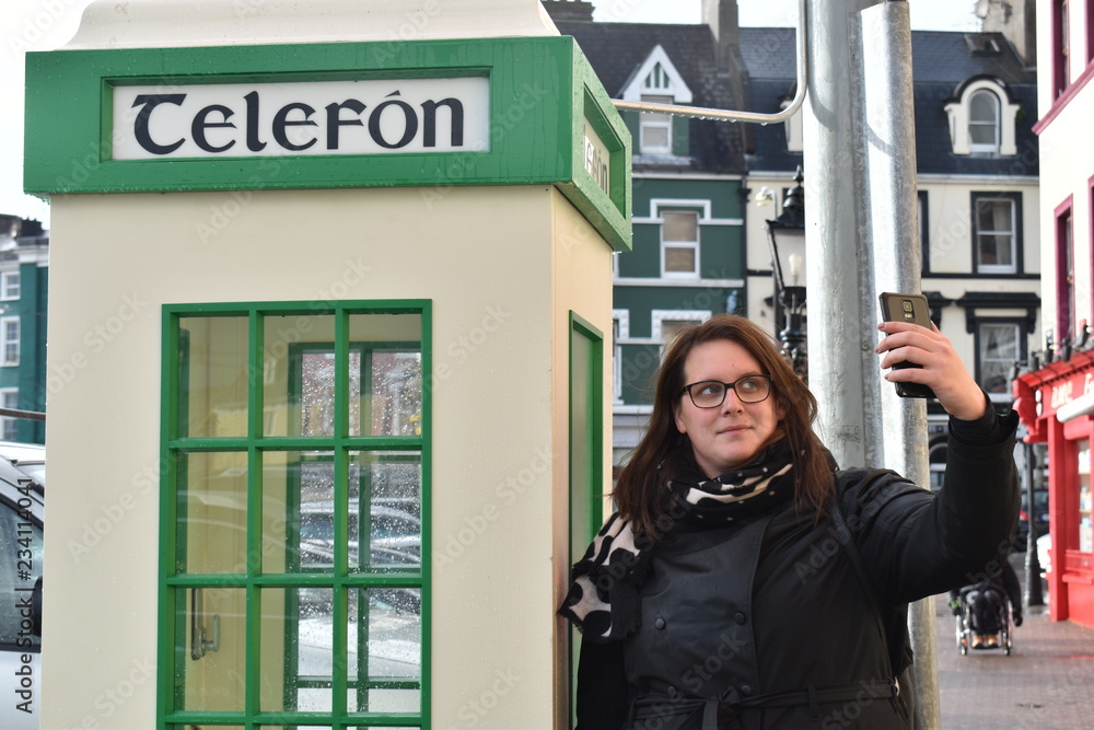 Lady in black taking a selfie with an Irish telephone box. The alternative to the red British telephone booth. Cobh, Ireland