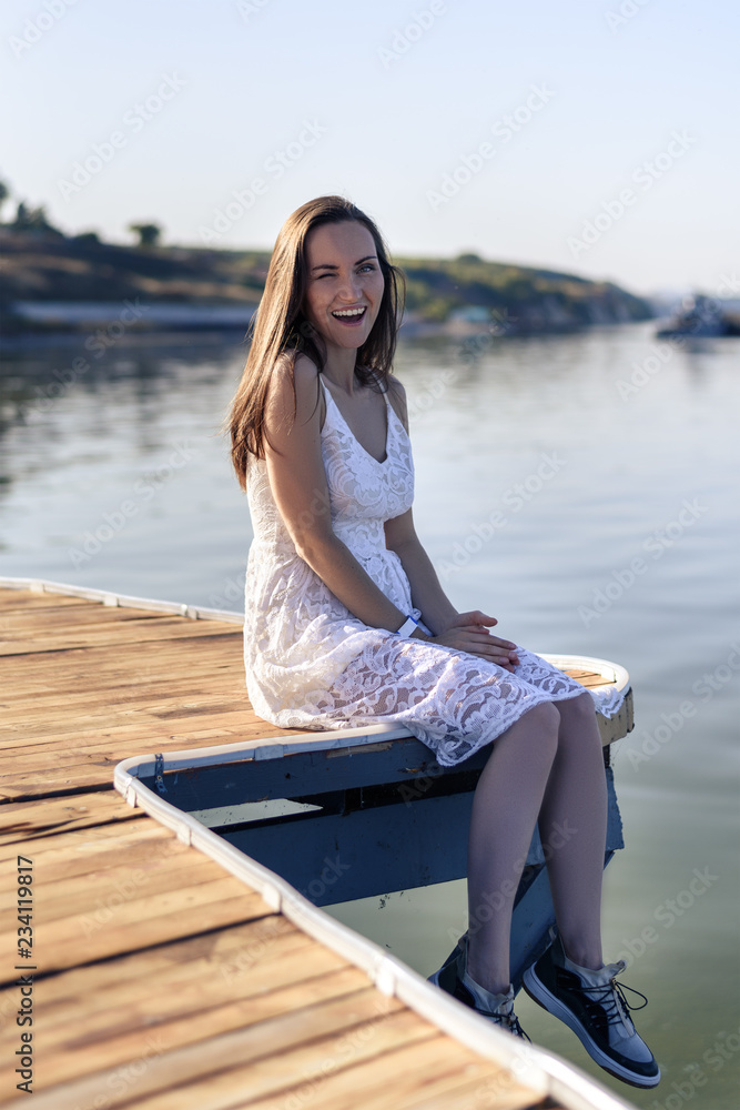 Young smiling woman winks her eye, sitting on the pier in a white sundress
