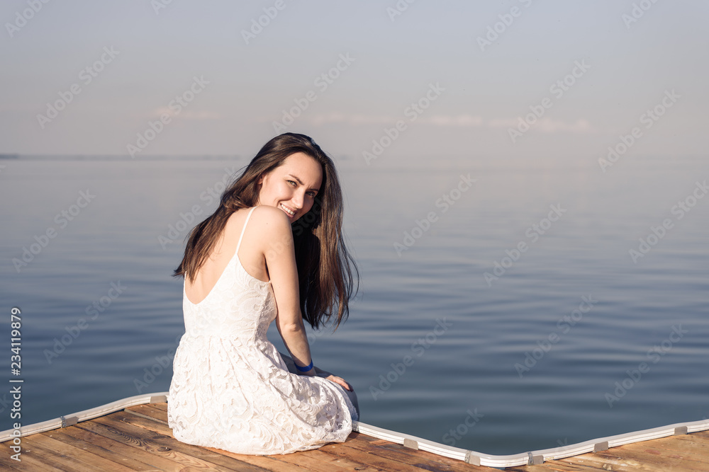 Woman sitting on the pier in half turn smiling in white dress on the background of blue sea