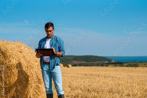 Front view of a male agronomist farmer with a tablet in blue jeans and a shirt and a white t-shirt in a field with haystacks © Руслан Галиуллин