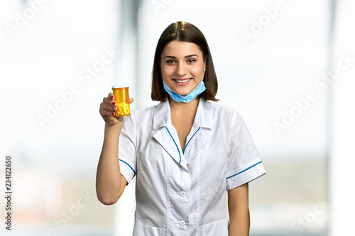 Woman doctor holding container with pills. Beautiful caucasian doctor in coat and mask holding orange bottle with medicaments.