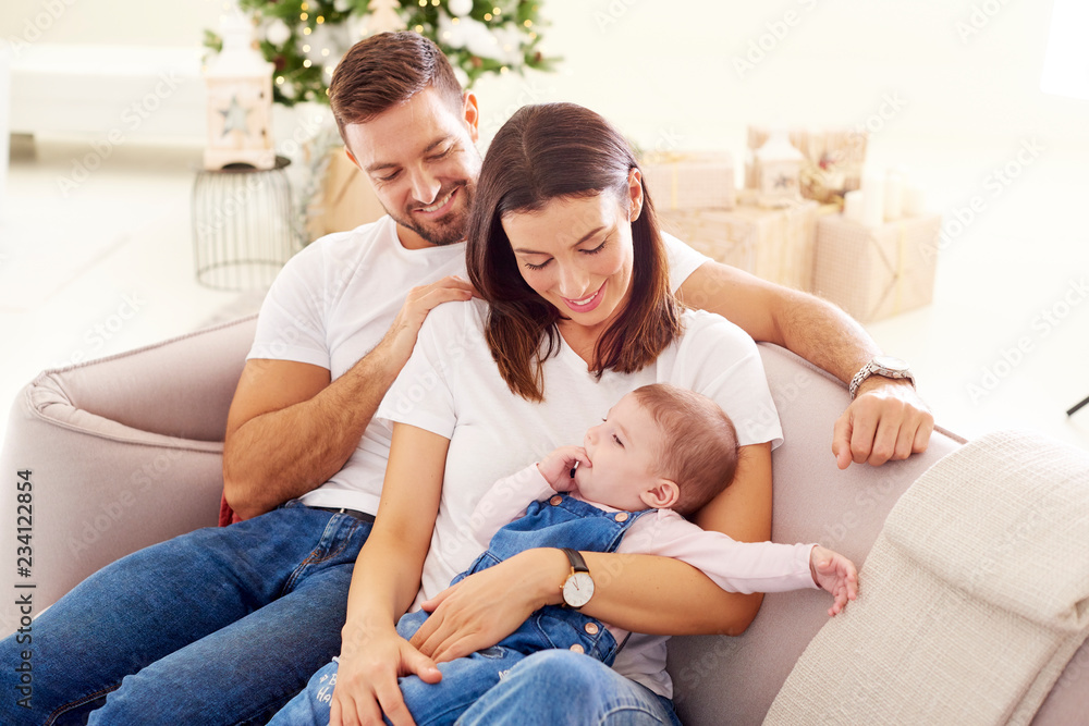 Happy young parents with their baby girl enjoy Christmas time while relaxing on the sofa. Family life.