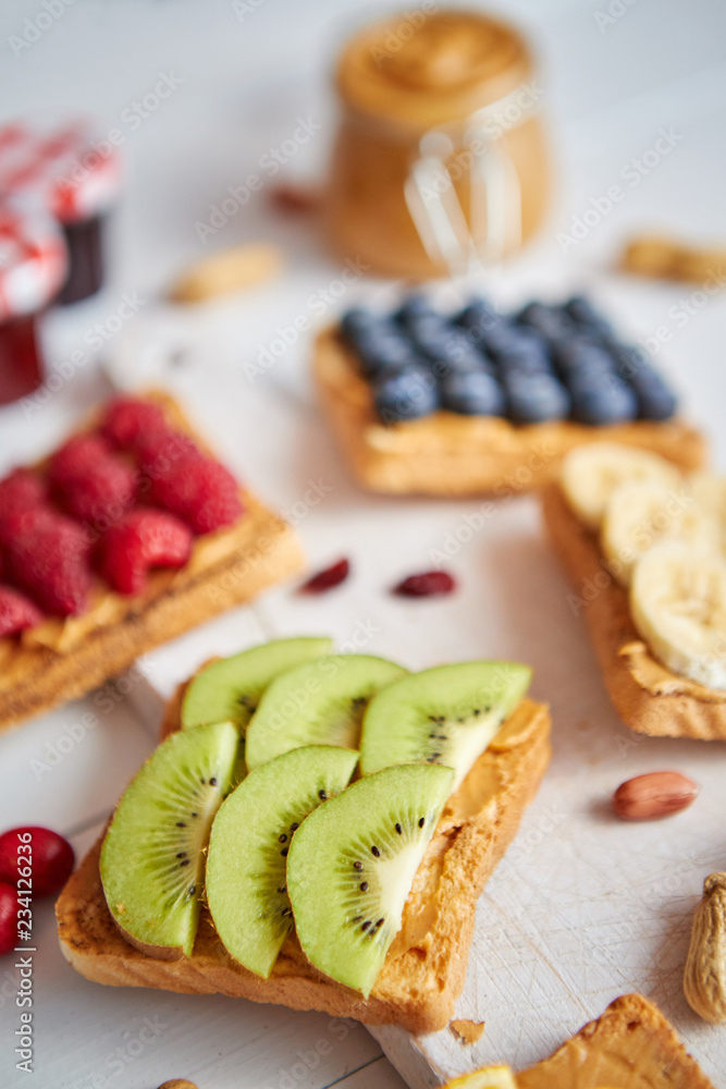 Assortment of healthy fresh breakfast toasts. Bread slices with peanut butter and various fruits and ingredients on side. Placed on white wooden table. Top view, with copy space. Selective focus Assortment of healthy fresh breakfast toasts. Bread slices with peanut butter and various fruits and ingredients on side. Placed on white wooden table. Top view, with copy space. Selective focus