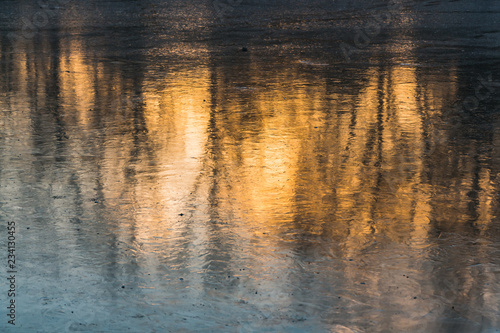 A beautiful texture of a pond with reflections of orange sunset and black trees on the transparent ice