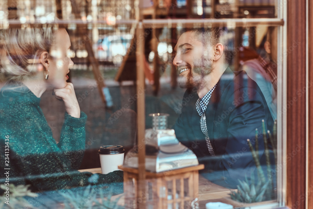 Attractive young couple looking at each other and talking while sitting in the restaurant behind the window. Attractive young couple looking at each other and talking while sitting in the restaurant behind the window.