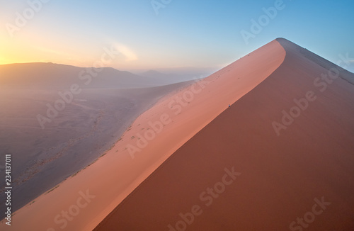 Aerial, artistic photo of dune with climber on its edge. Early morning Namib desert covered in mist. Orange dunes of Namib from above. Desert landscape. Sunrise in Namib desert. Traveling Namibia. © Martin Mecnarowski