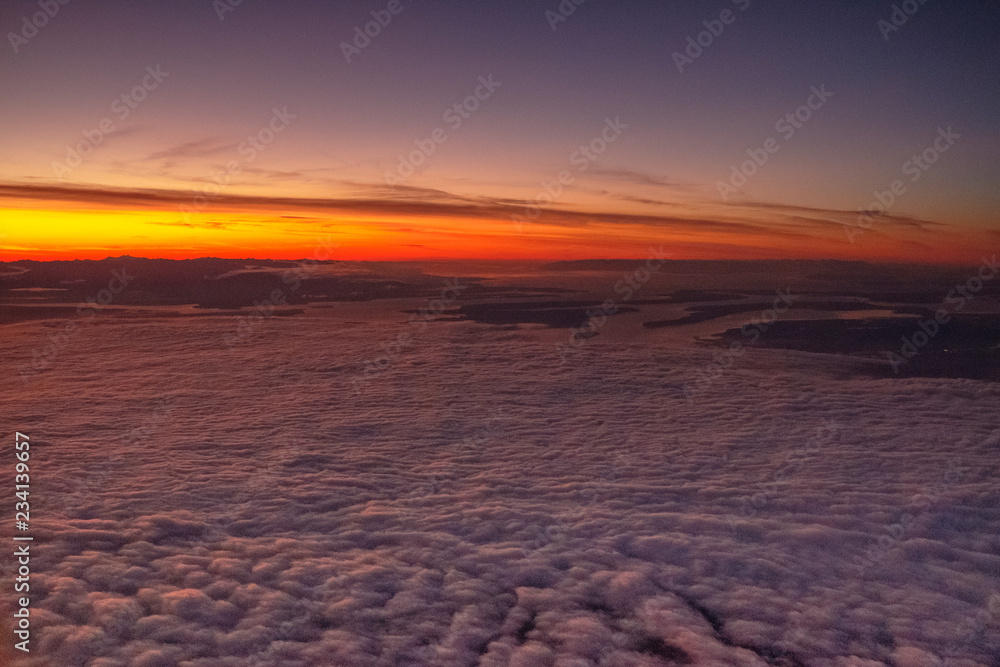 The golden sun casts its rays across the tops of clouds at sunset while lit cities await below
