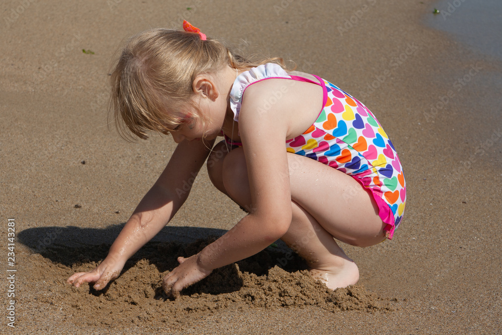 Little girl in a colorful bikini playing with sand on the sea beach ...