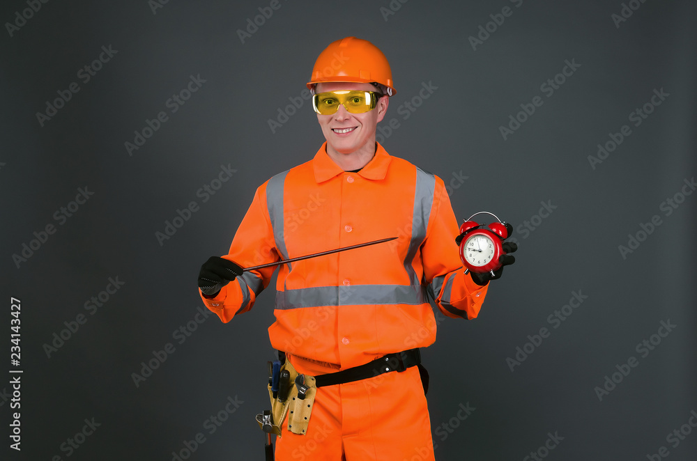 Happy builder worker is showing on a red alarm clock in his hand isolated on gray background. Time to work. Building construction time. Happy builder worker is showing on a red alarm clock in his hand isolated on gray background. Time to work. Building construction time.