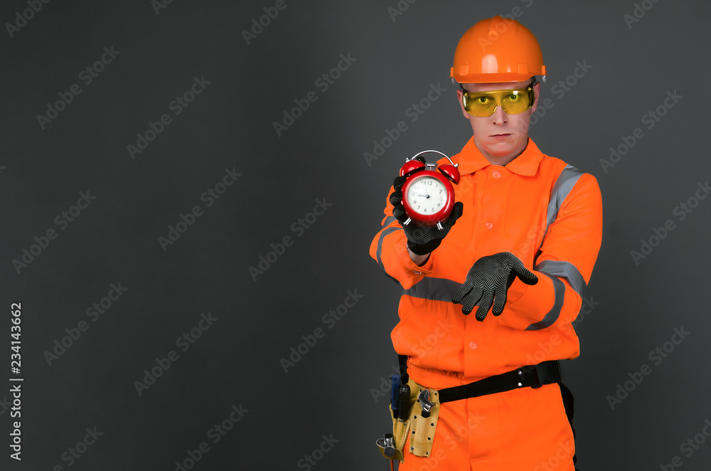 Angry builder foreman in the hardhat with red alarm clock in the hands isolated on gray background with copy space. Lateness in the construction schedule plan of the housing object concept. Angry builder foreman in the hardhat with red alarm clock in the hands isolated on gray background with copy space. Lateness in the construction schedule plan of the housing object concept.