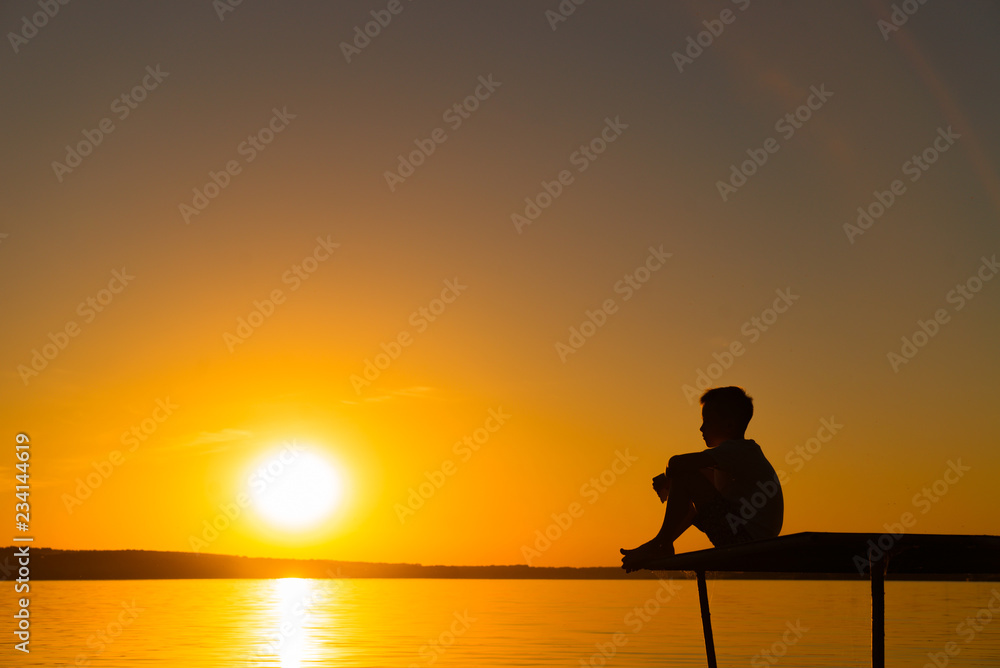 Silhouette of a little boy sitting on the bridge in river at the sky sunset. A lonely boy sits quietly and looks ahead at a beautiful view of sunset.