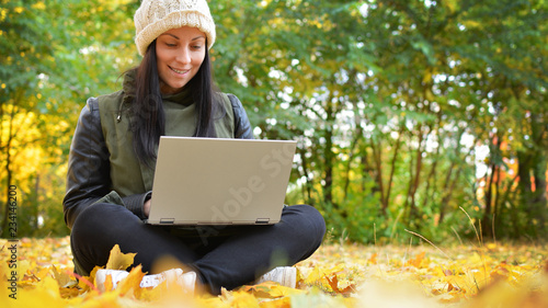 Girl in hipster with laptop in autumn park. A woman in a cap using a laptop while sitting on fallen leaves. Freelancer in the hat uses remote communication technology. Remote work. Free space