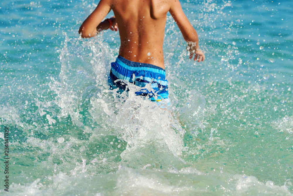 Cute European boy in colorful swimming shorts is running towards the ocean, making numerous splashes around. He is happy to spend his summer holidays at the sea cost.
